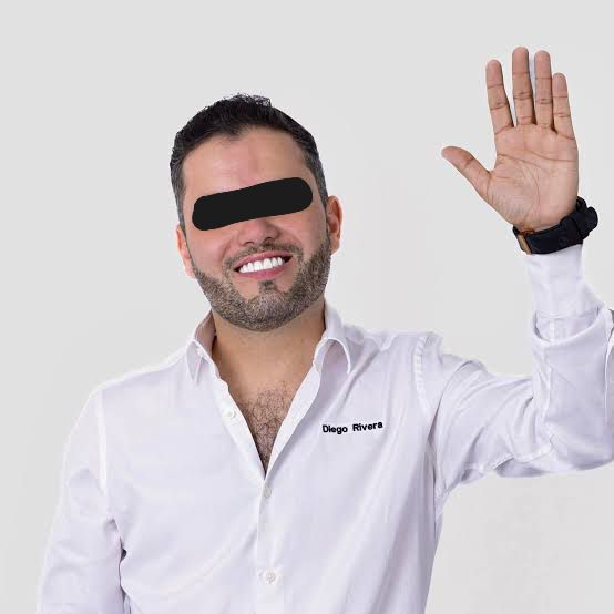 A man with a beard and short hair wearing a white shirt, smiling and waving with his right hand.