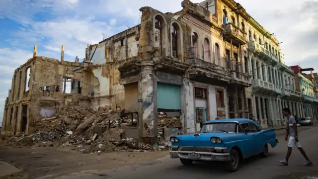 A vintage blue car parked on a street in Havana, Cuba, with a backdrop of a partially collapsed building and historical architecture.
