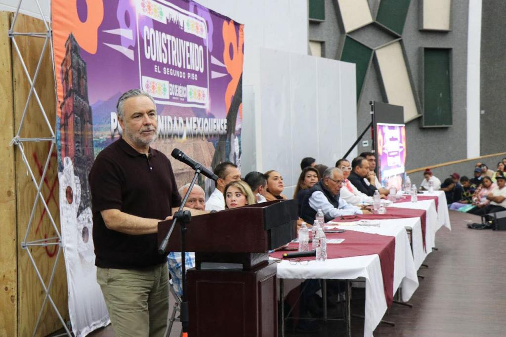 Un orador en un podio presenta un evento en una sala con un fondo decorado, mientras una audiencia observa desde un lado.