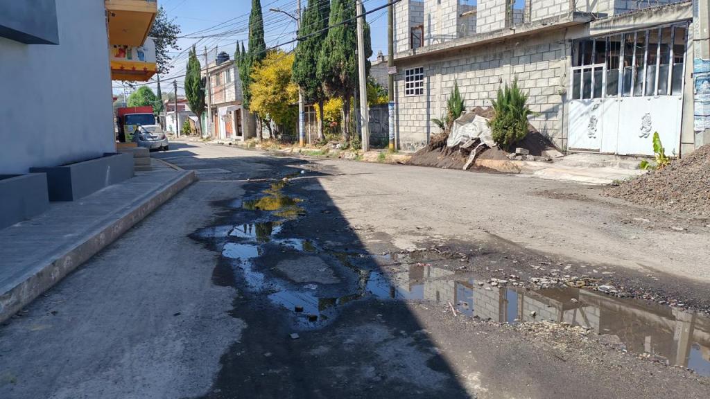 A street view showing a damaged road with puddles, surrounded by residential buildings and trees.