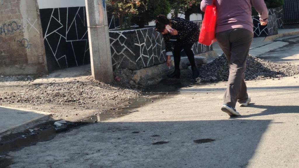 A street scene showing two people: one is bending down next to a wall, possibly fixing something, while another person is walking by carrying a red bag. The ground is uneven with gravel and some water pooling on the street.
