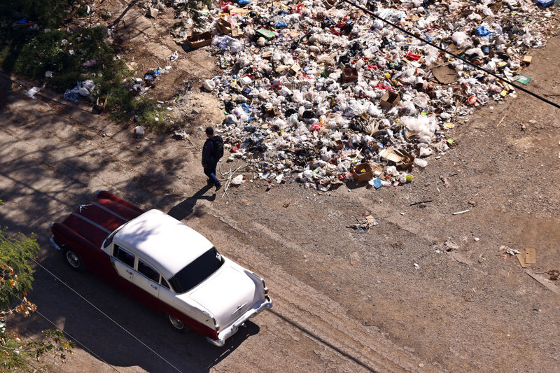 Un automóvil antiguo estacionado cerca de una acumulación de basura en un terreno árido, con una persona caminando al lado.
