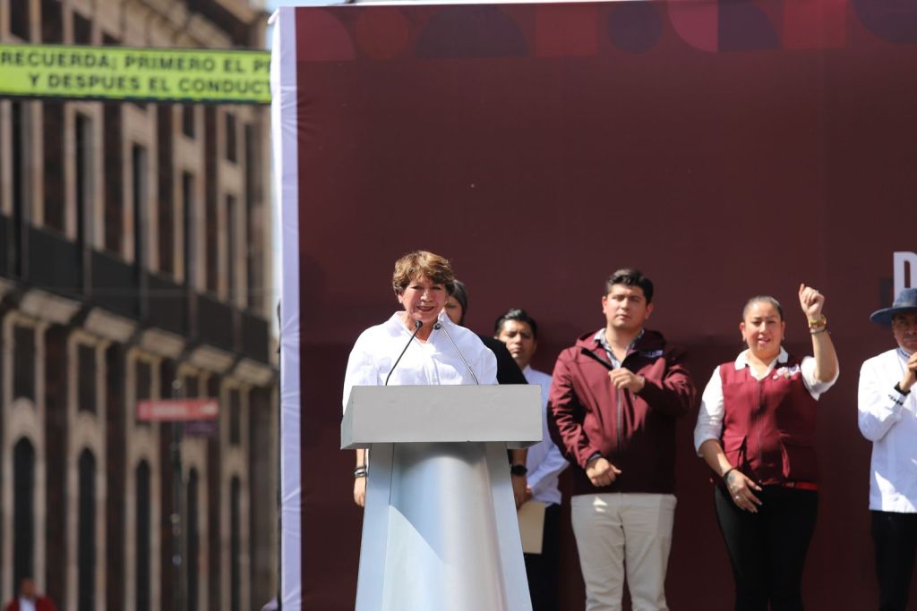 A speaker stands at a podium addressing an audience, with several supporters visible behind. The setting appears to be an outdoor event with a backdrop featuring a maroon design.