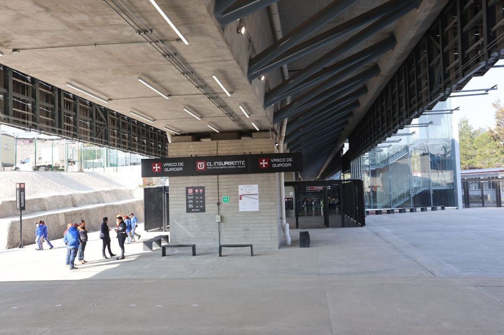 Entrance to a modern transport station with a concrete overhang, signage for 'Vasco de Quiroga' and 'El Insurgente', and people walking in front.