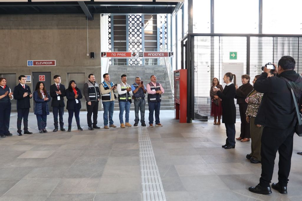 A group of people gathered in a modern building interior, some clapping and others observing, with signs indicating 'Elevador' and 'No Pase' in the background.