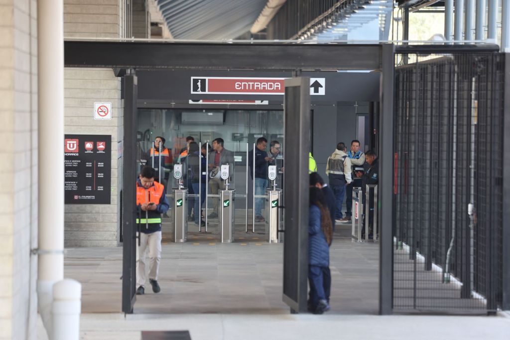 Entrance area of a transportation facility with people passing through turnstiles and a sign indicating 'Entrada'. Various individuals are seen, some in uniforms, and a list of service hours is displayed on a wall.
