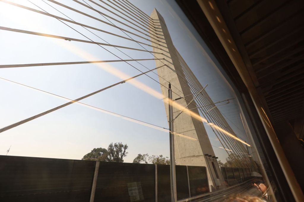View of a tall concrete bridge pylon with cables against a clear sky, seen through a train window.