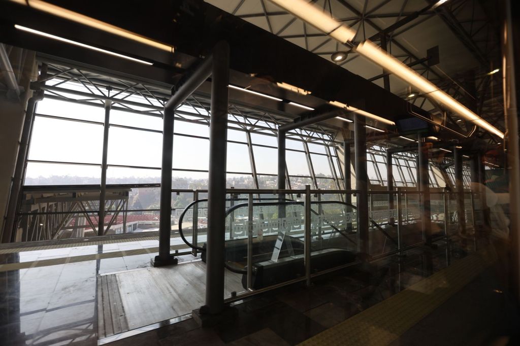 Interior view of a modern train station platform with large glass windows and an escalator, overlooking a cityscape.