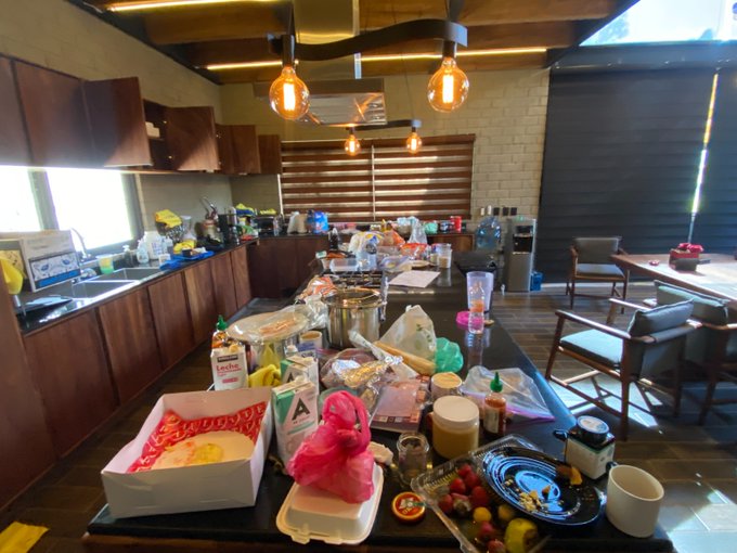Cocina desordenada con una amplia variedad de alimentos y utensilios en la encimera, iluminada por lámparas colgantes. Hay sillas y una mesa al fondo.