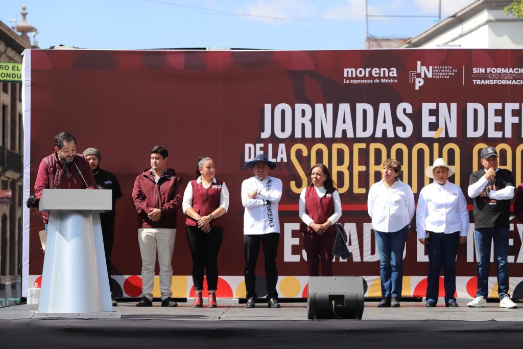 A group of politicians on stage during an event titled 'Jornadas en Defensa de la Soberanía' in Mexico, with a speaker at a podium and attendees dressed in various colored attire.