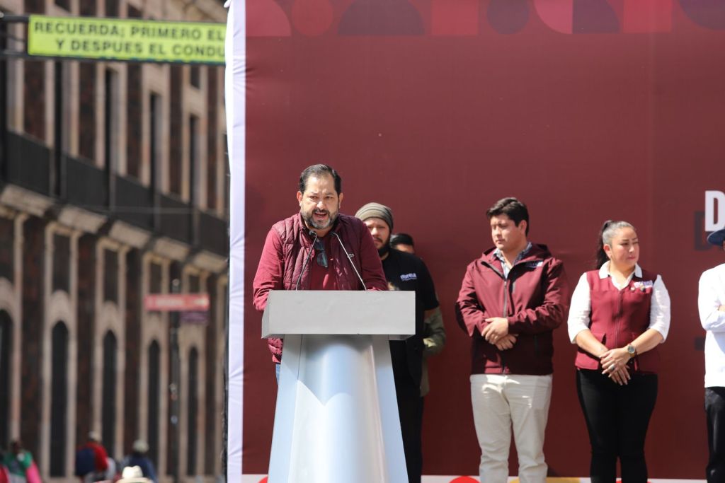 A speaker at a podium addressing an audience, with several people standing behind him, in a public event setting.
