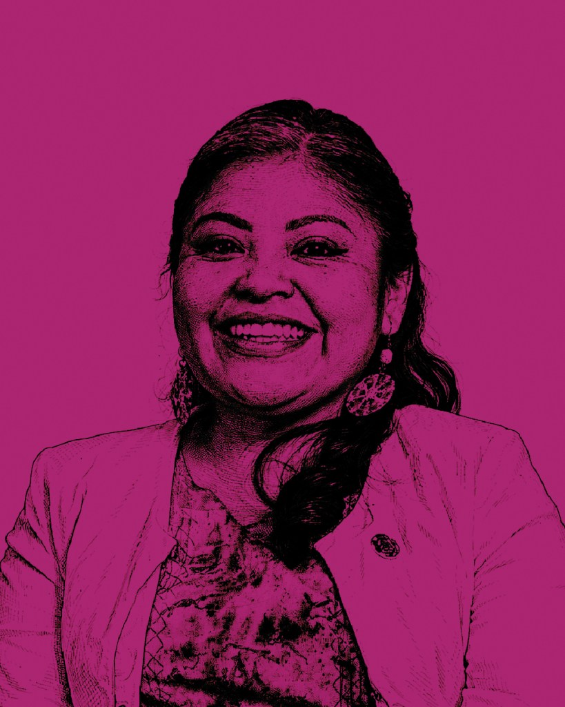 Retrato de una mujer sonriendo, con cabello oscuro y pendientes grandes, vestida con una chaqueta blanca y un top colorido, sobre un fondo púrpura.