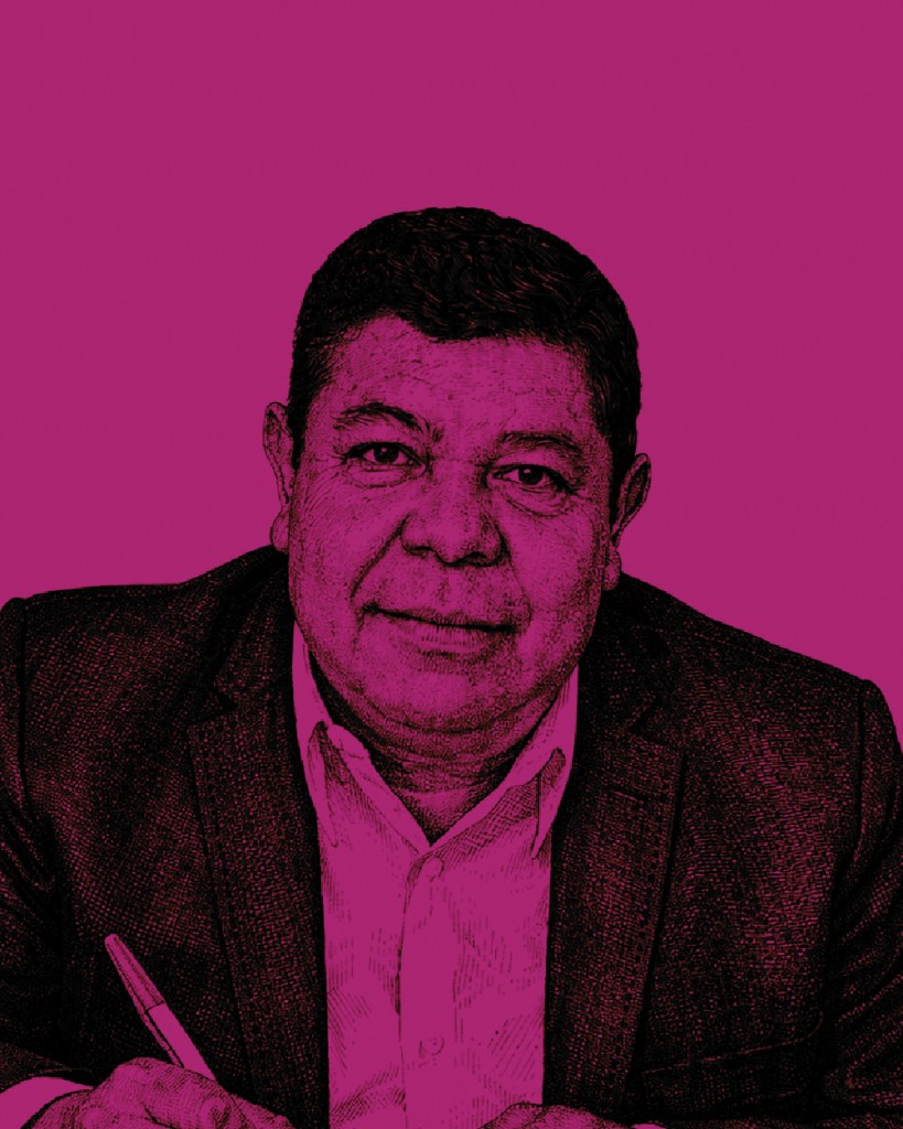 Retrato de un hombre con chaqueta oscura y camisa clara sobre un fondo rosa. Está sentado y sosteniendo un bolígrafo.