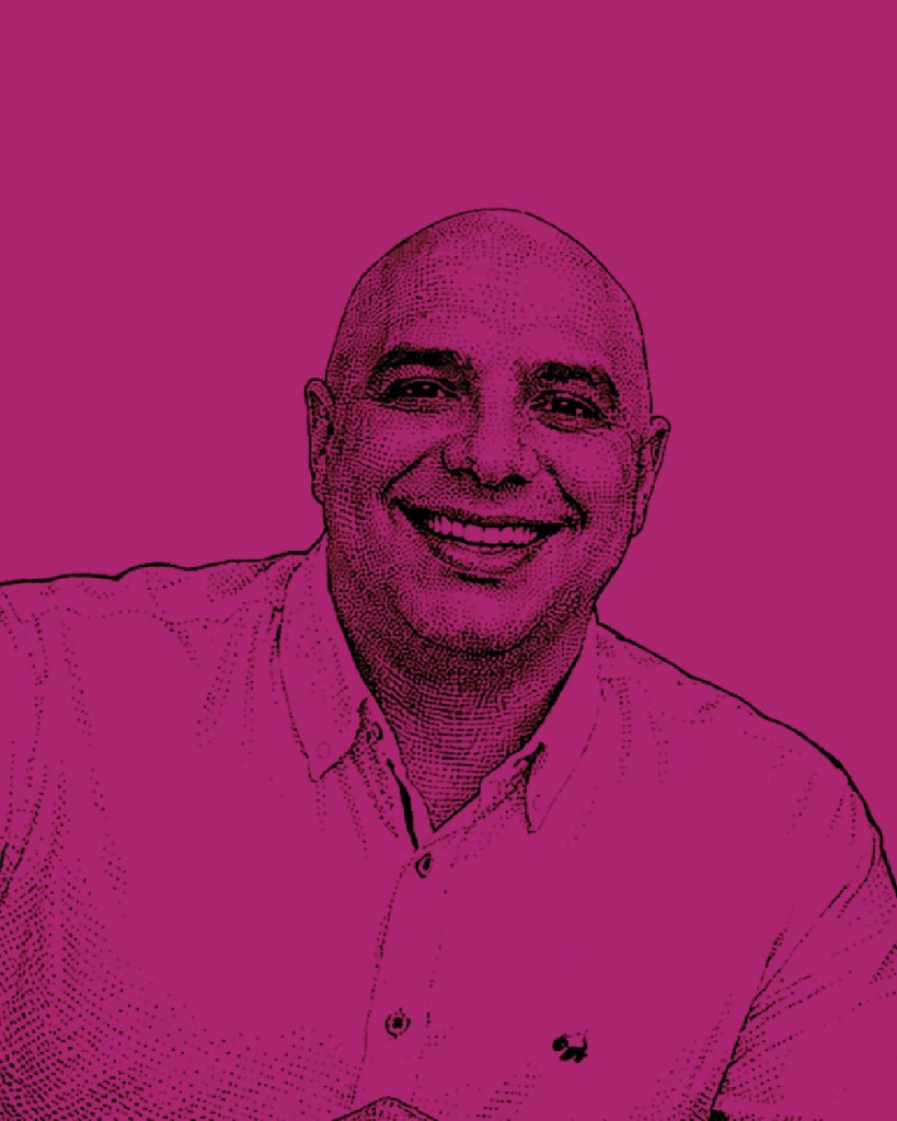 Retrato de un hombre sonriente con cabeza rapada, vestido con una camisa de botones blanca, sobre un fondo rosa.