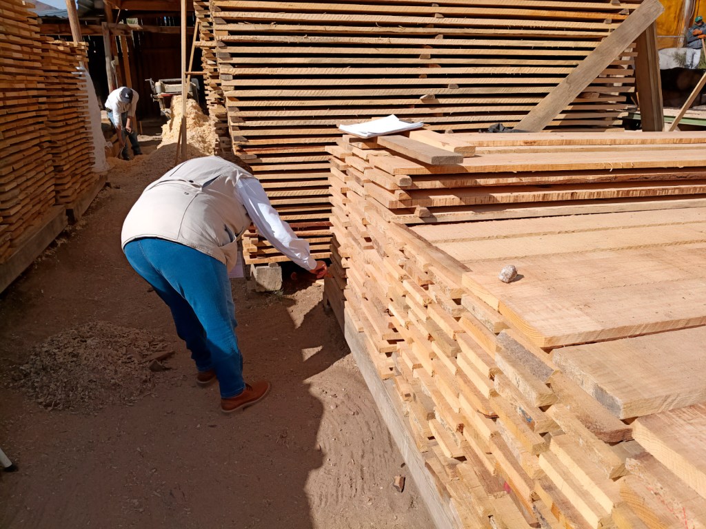 A worker inspecting stacked wooden planks in a lumber yard, with another worker digging in the background.