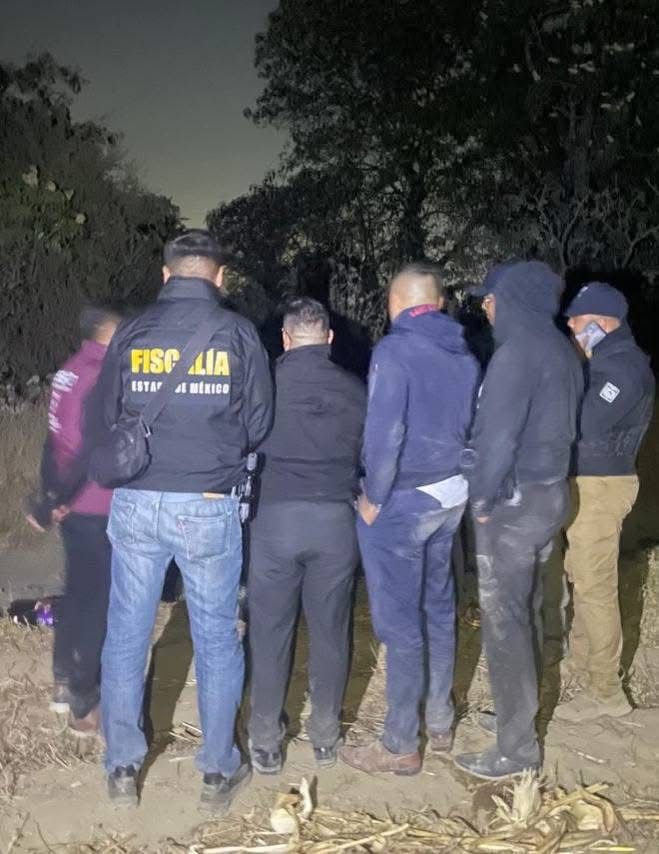 A group of individuals, some in law enforcement attire, standing in a dimly lit area surrounded by trees and vegetation.