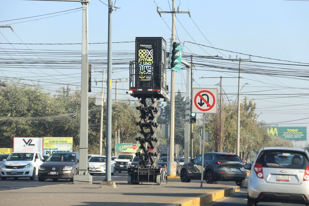 A traffic signal and a mobile advertising unit elevated on a scissor lift, positioned at an intersection with vehicles passing by. Nearby traffic signs indicate regulations, and overhead power lines are visible.