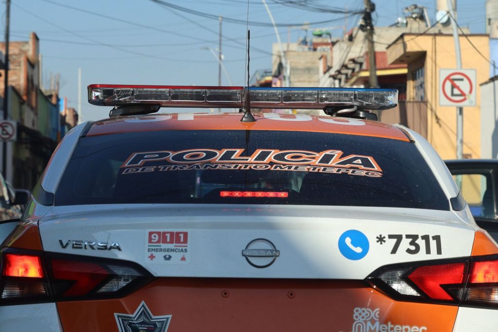 Rear view of a police car displaying 'Policía de Tránsito Metepec' with emergency contact information and a warning light on top, parked in an urban setting.