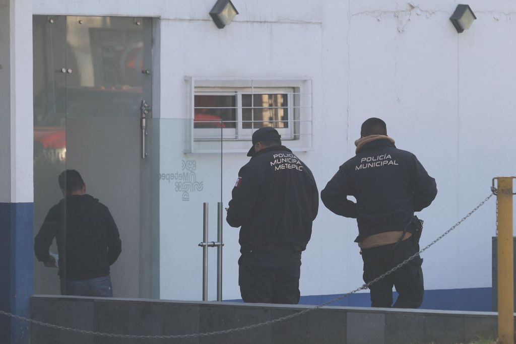 Two police officers in dark jackets with 'Policía Municipal' written on them stand outside a building, while a third person in a black coat walks away from them.
