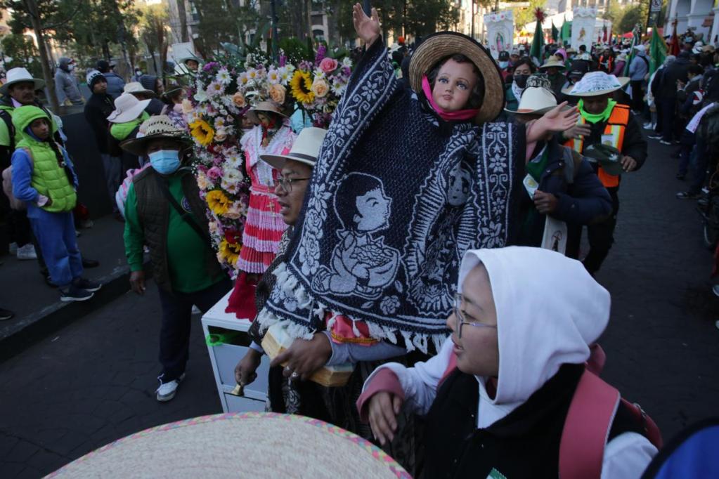 Una multitud celebra un evento cultural con personas vestidas en trajes tradicionales. Una figura artística similar a una muñeca, vestida con un poncho y que levanta los brazos, es el foco central, rodeada de flores y varios asistentes que llevan sombreros.