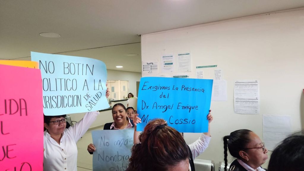 Protesters holding signs in an office setting, demanding the presence of Dr. Angel Enrique Cossio and opposing political interference in health jurisdiction.