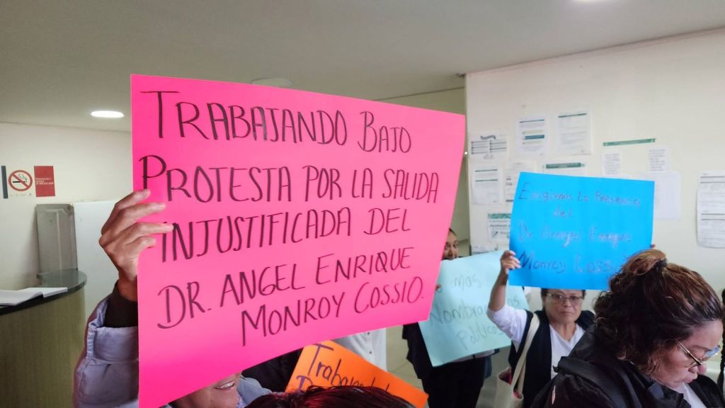 Protesters holding colorful signs demanding the unjustified removal of Dr. Angel Enrique Monroy Cossio, inside a building.