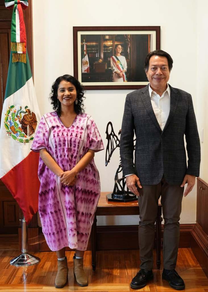 A woman in a colorful traditional dress stands next to a man in a blazer and white shirt, smiling in front of a Mexican flag and a portrait in a formal setting.
