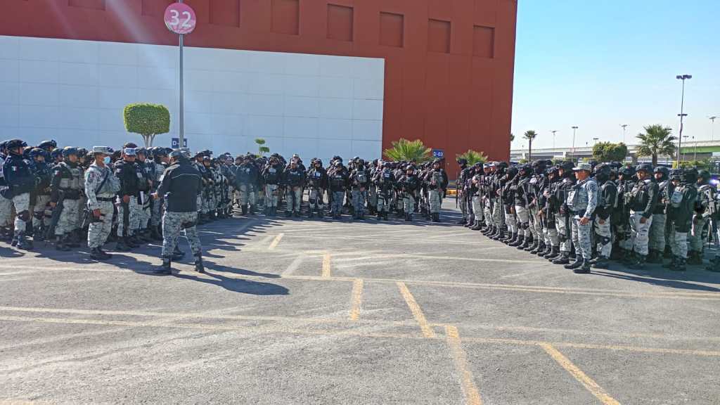 A large group of uniformed police officers standing in formation, listening to a speaker, in a parking area with a building in the background.