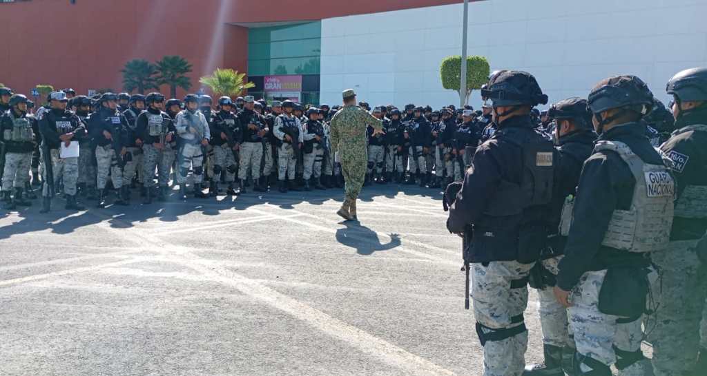 A uniformed officer addresses a large group of armed personnel gathered in a training or briefing setting outside a building.
