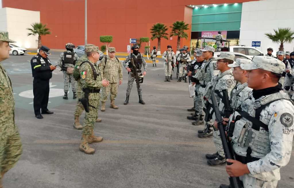A group of military and police personnel gathered in a parking area, some in uniform discussing and others standing in formation with weapons at the ready.