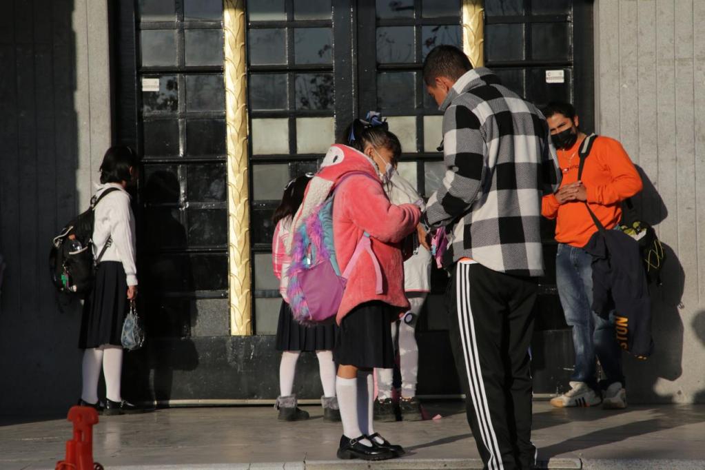 A group of school children and adults gather outside a building, with some children wearing uniforms and backpacks, while adults are engaged in conversation.