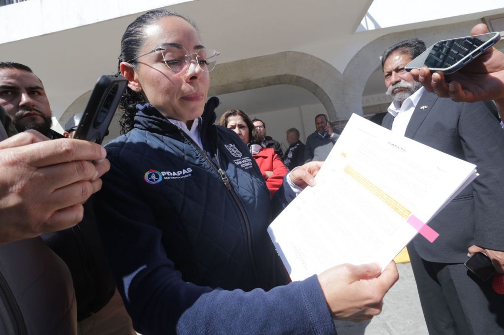 Una mujer con gafas sostiene un documento mientras es grabada por un teléfono móvil. A su alrededor, un grupo de personas observa atentamente.