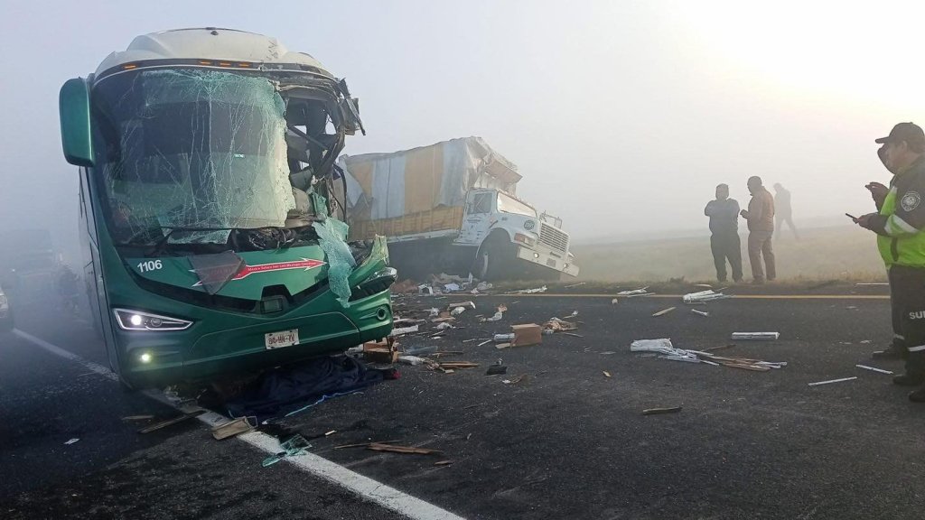 Image of a severe bus accident on a foggy highway, featuring a damaged green bus with shattered windows and debris scattered around, while several people and emergency responders assess the scene.