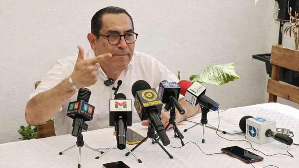 Hombre con gafas y camisa blanca gesticulando en una conferencia de prensa, frente a varios micrófonos de medios de comunicación.