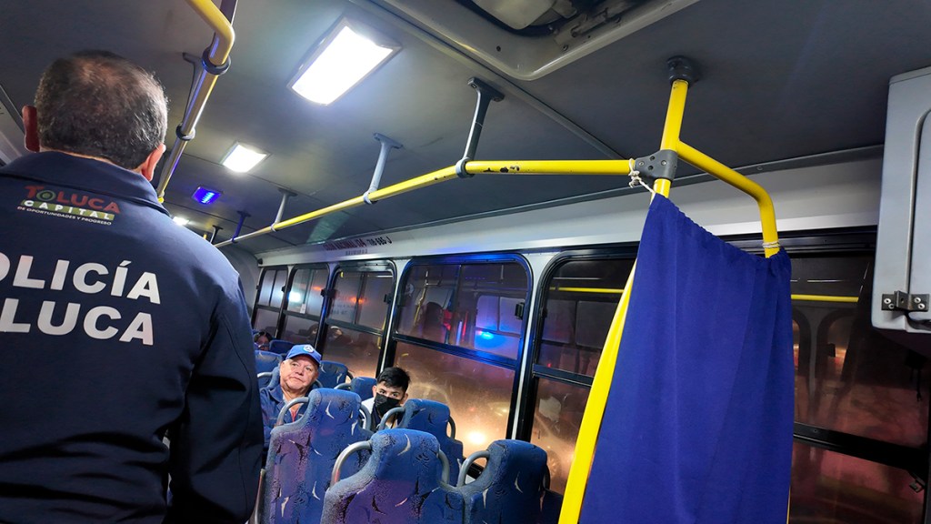 Interior de un autobús con un oficial de policía de espaldas, mientras dos pasajeros lo observan. La iluminación es tenue y hay una cortina azul en el fondo.