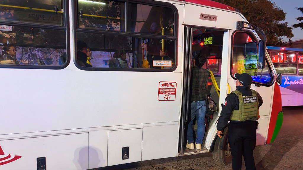 Un autobús de transporte público con pasajeros subiendo y bajando, mientras un policía municipal observa la situación.