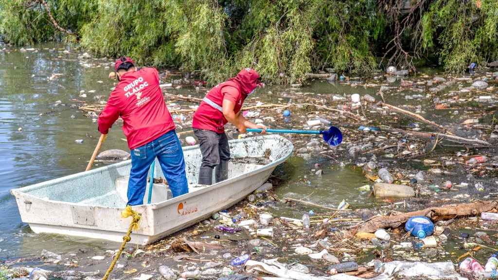 Two individuals in red shirts are cleaning a polluted waterway while using tools to remove debris from the water. The scene shows a significant amount of trash and plastic waste floating in the water and along the banks.