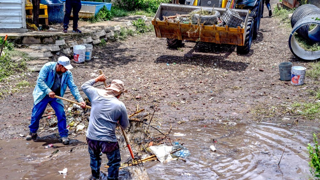 Two men working in a muddy area, using tools to remove debris, while a tractor is seen in the background transporting waste.