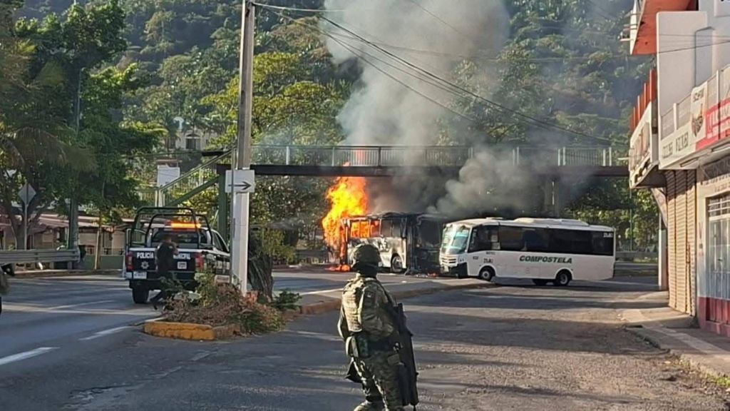 Un autobús en llamas en una calle, con humo denso saliendo y un soldado de pie en el primer plano, observando la escena. Al fondo, otro autobús y un vehículo policial.