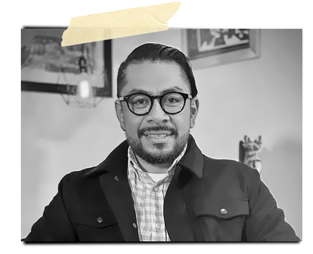 Retrato en blanco y negro de un hombre con gafas, sonriendo y vistiendo una chaqueta oscura sobre una camisa a cuadros. Se observa un entorno decorativo con fotografías en la pared.