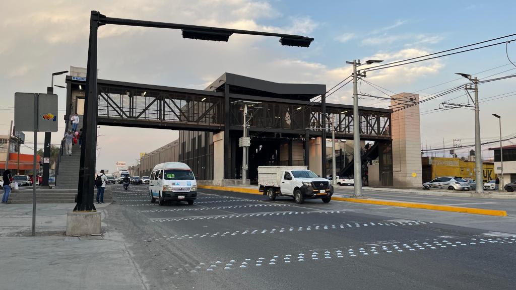 Vista de una estación de transporte urbano con un puente peatonal, vehículos en la calle y tráfico en movimiento.