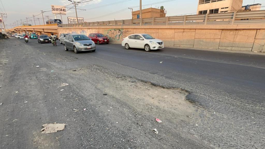 Vista de una calle con tráfico vehicular, donde se observa un bache en la carretera y un entorno urbano con basura a los lados.