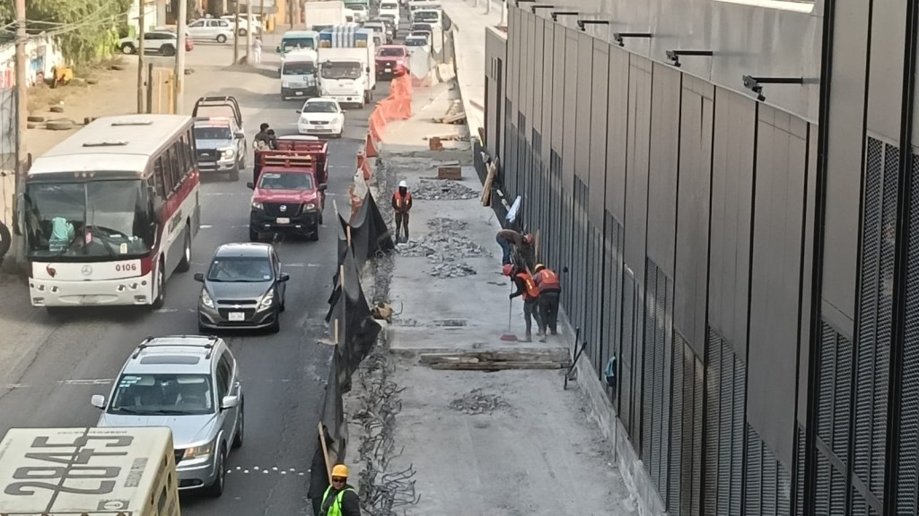 Imagen que muestra una calle en construcción con varias camionetas y automóviles en movimiento, mientras trabajadores con chalecos naranjas realizan labores de construcción en la acera.