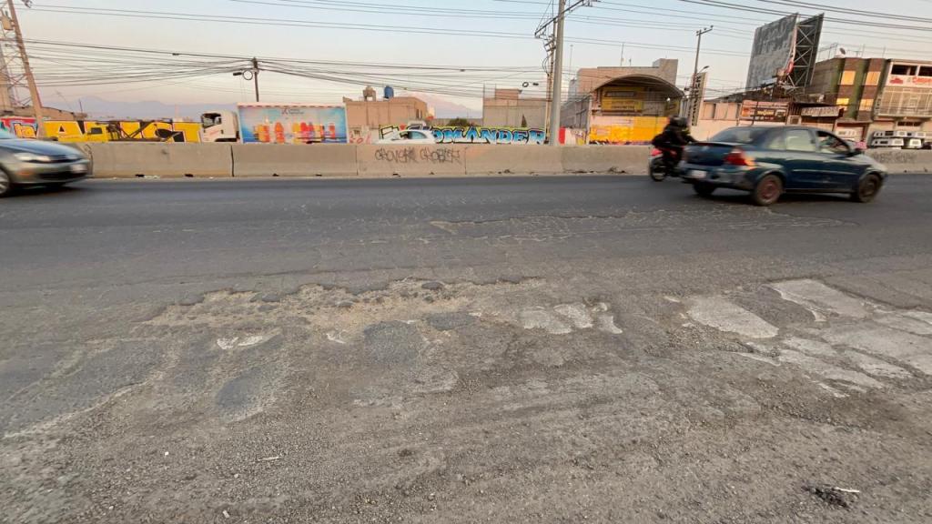 Vista de una carretera con pavimento dañado y vehículos en movimiento, incluyendo un automóvil y una moto.