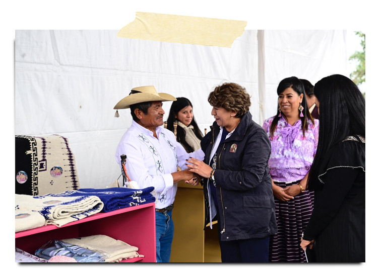 Un hombre con sombrero y una camisa blanca se está saludando con una mujer en un ambiente festivo. Detrás, hay varias personas observando y productos artesanales en una mesa.