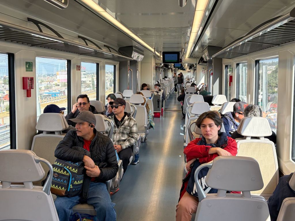 Interior of a train with several passengers seated, some engaged in conversation and others looking out the windows. The interior features modern design with bright lighting and visible advertisements outside.