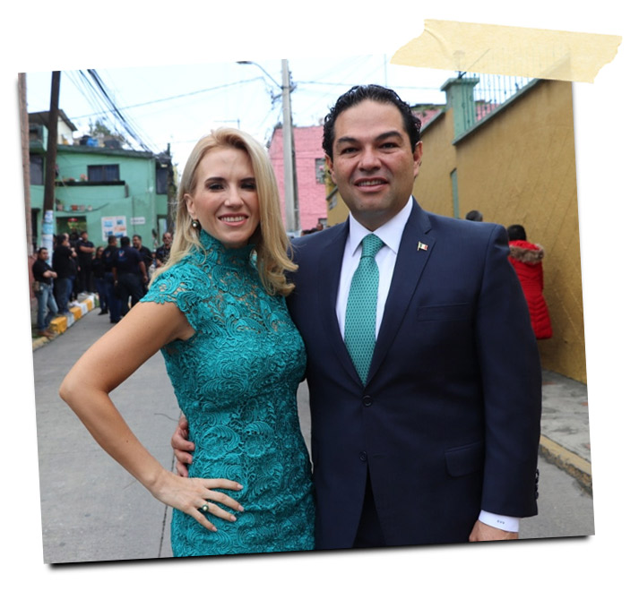 Una mujer con un vestido verde y un hombre con traje oscuro y corbata verde posan juntos sonrientes en una calle, rodeados de edificios coloridos.
