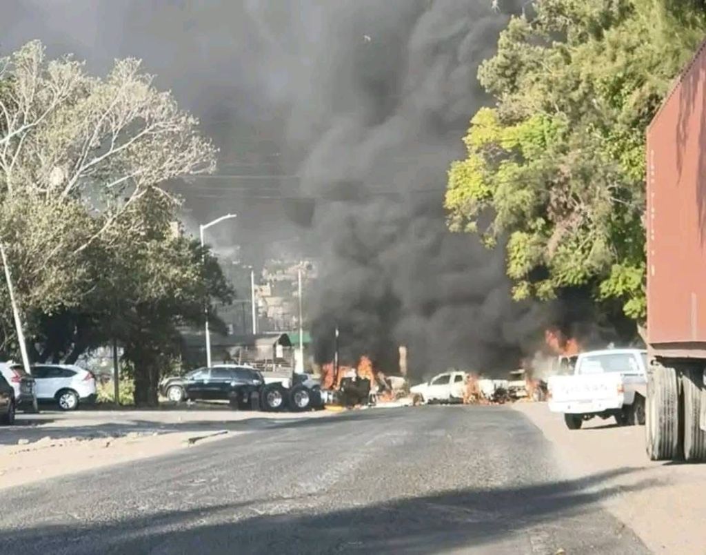 Una carretera con vehículos en llamas y una densa columna de humo negro en el fondo.