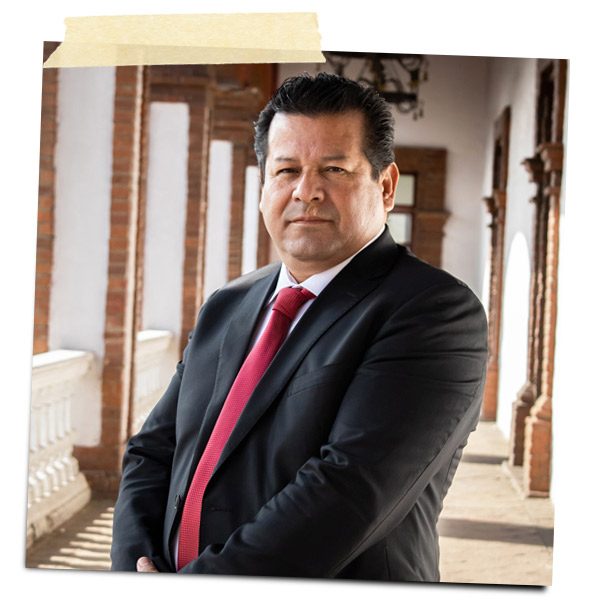 Hombre de pie con traje negro y corbata roja, posando en un entorno arquitectónico con paredes de ladrillo y arcos.