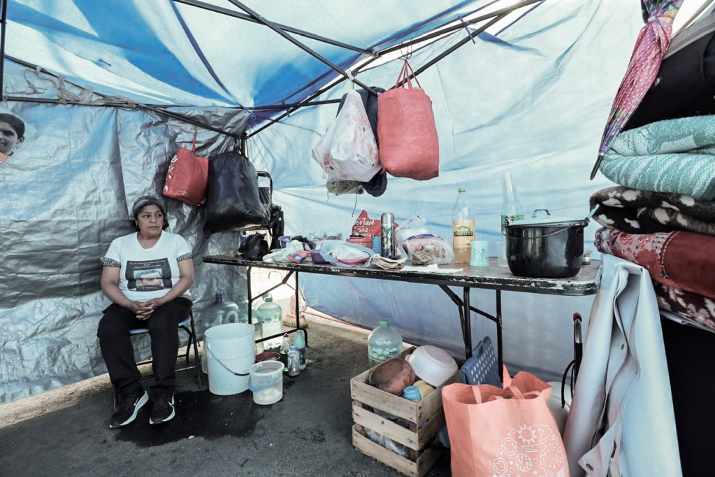 Mujer sentada en una tienda temporal, rodeada de bolsas, utensilios de cocina y alimentos sobre una mesa.